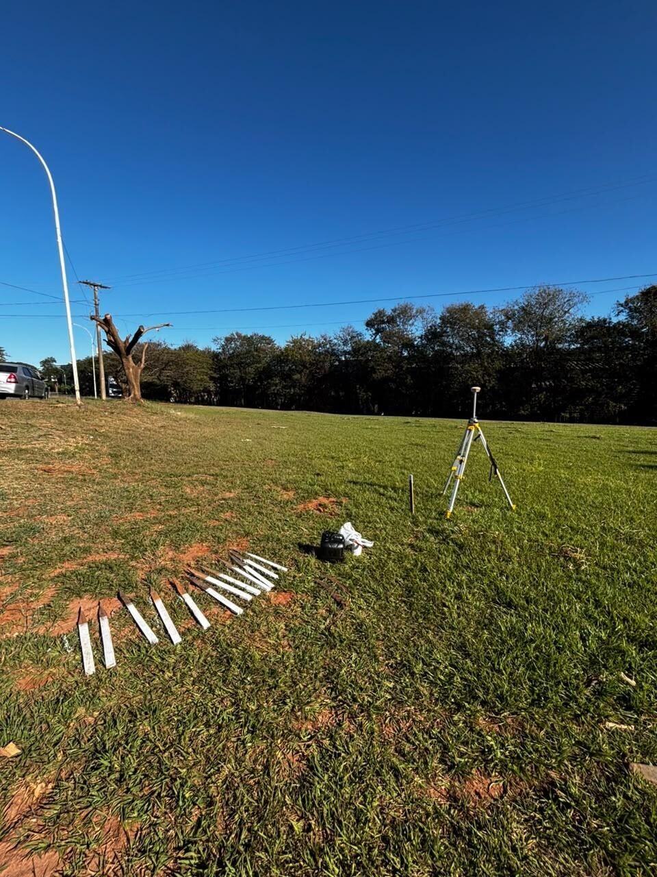 Estacas de madeira pintadas de branco enfileiradas na grama, ao lado de um receptor GNSS sobre tripé