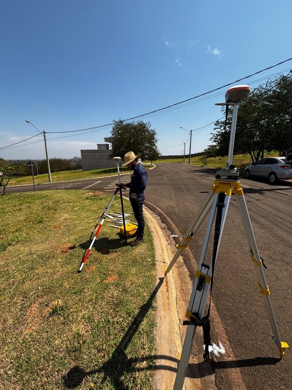 Receptor GNSS sobre tripé junto ao meio-fio de uma rua, com um técnico ao fundo