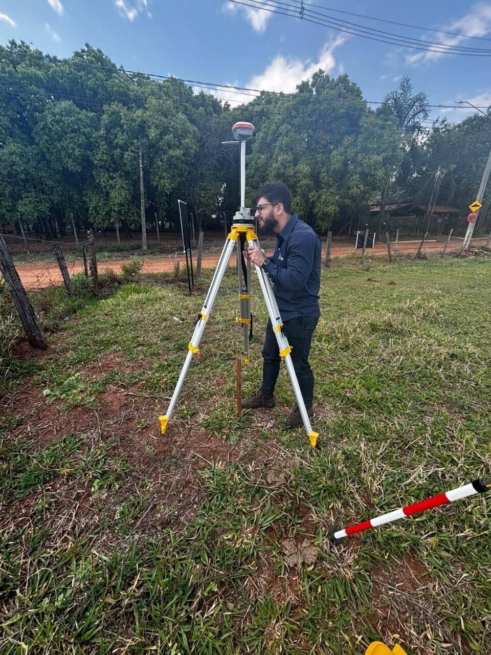 Técnico da RC Geo operando um receptor GNSS sobre tripé em terreno gramado, com árvores ao fundo