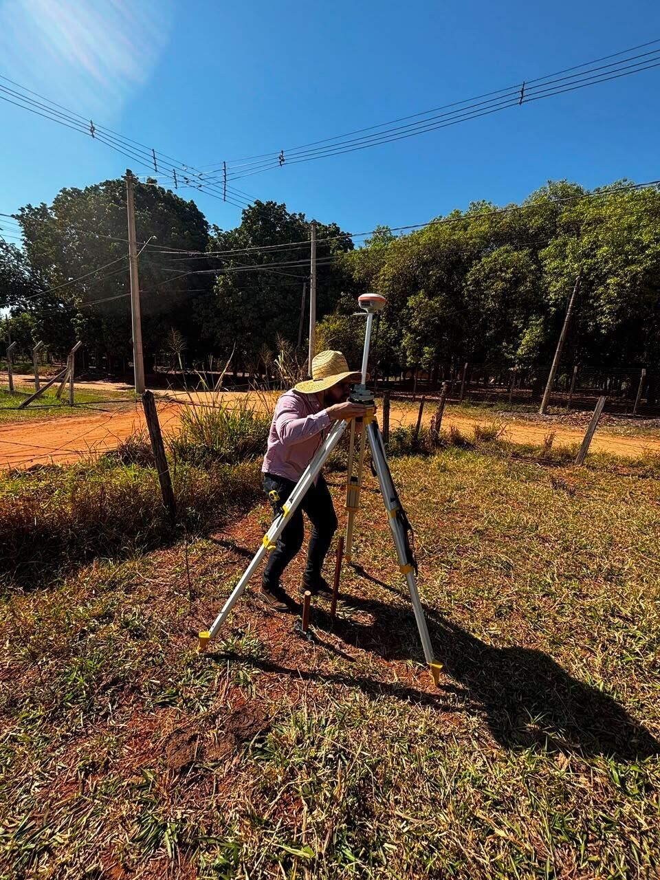 Técnico da RC Geo de chapéu de palha operando um receptor GNSS sobre tripé em estrada de terra