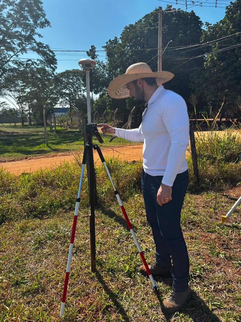 Técnico da RC Geo de chapéu de palha ajustando um receptor GNSS sobre tripé em campo aberto