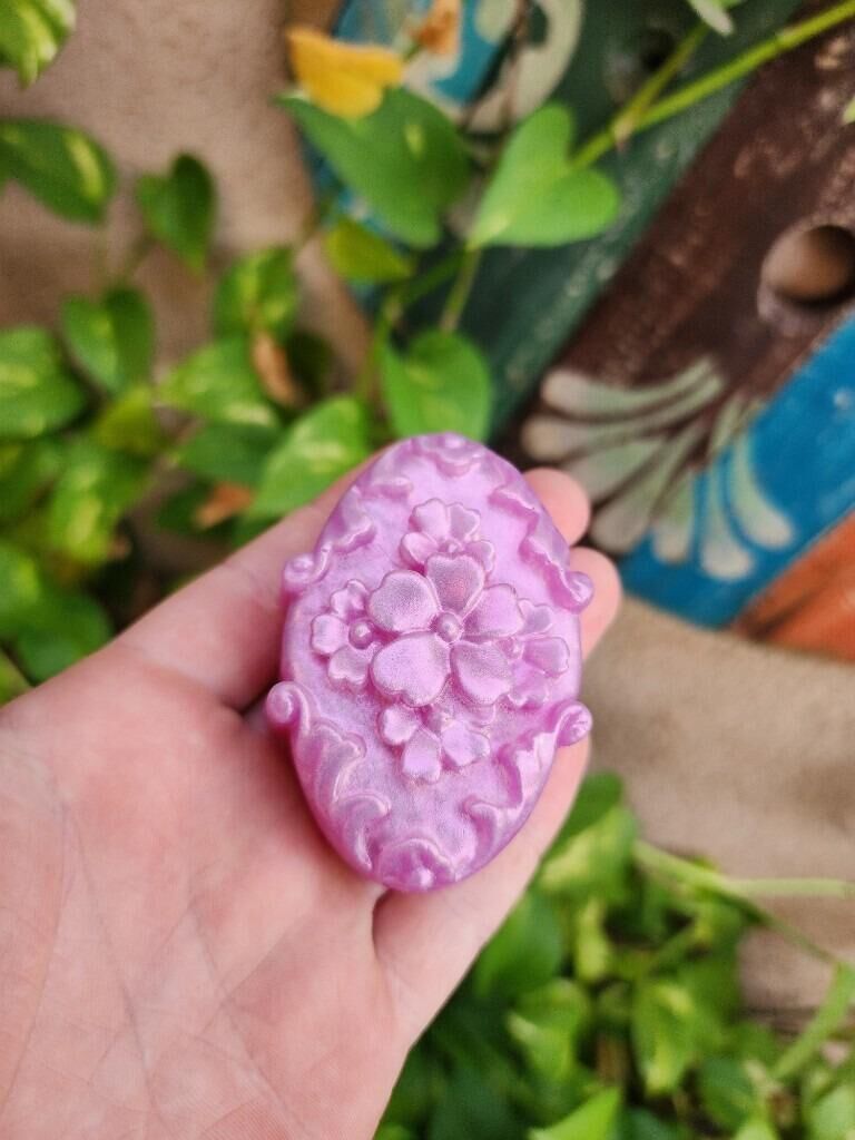 Sabonete Floral Lavanda, oval lilás com flor em relevo, na mão entre folhas
