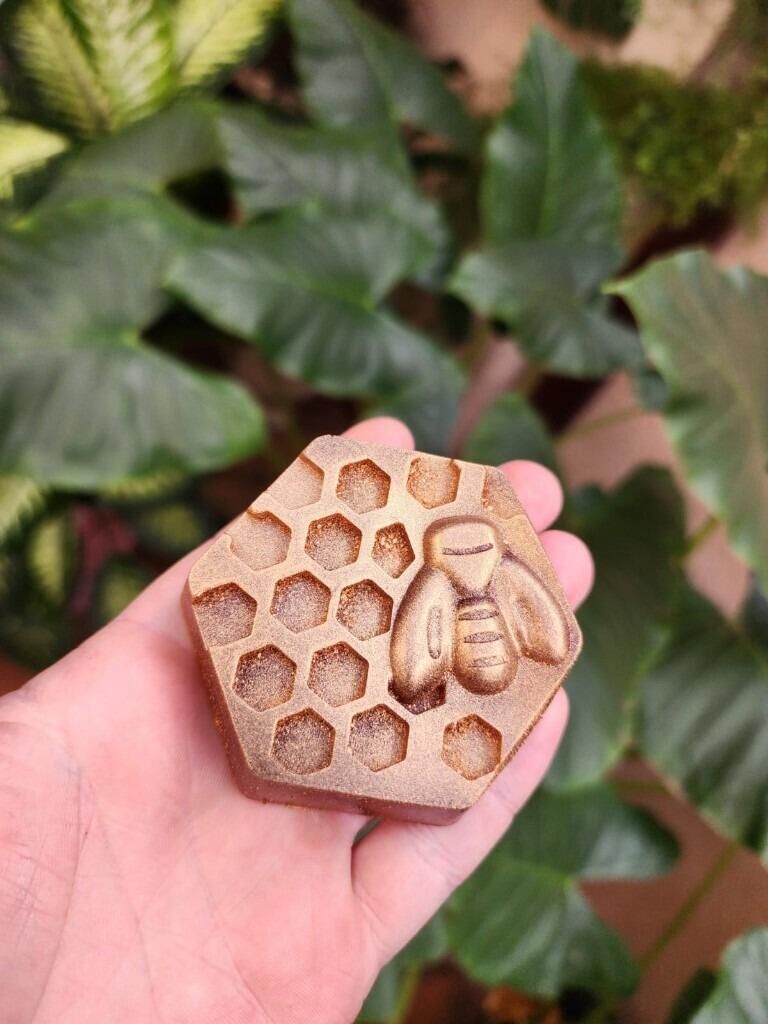 Mão segurando sabonete hexagonal dourado com favos e abelha em relevo